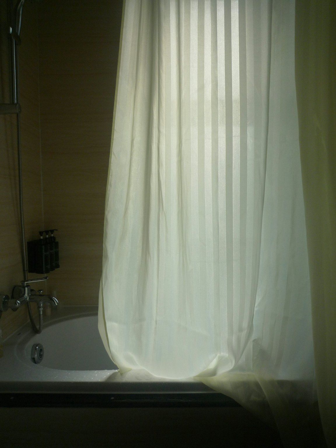 Un rideau de douche blanc à rayures tiré devant une baignoire, avec un flexible et une robinetterie à gauche.