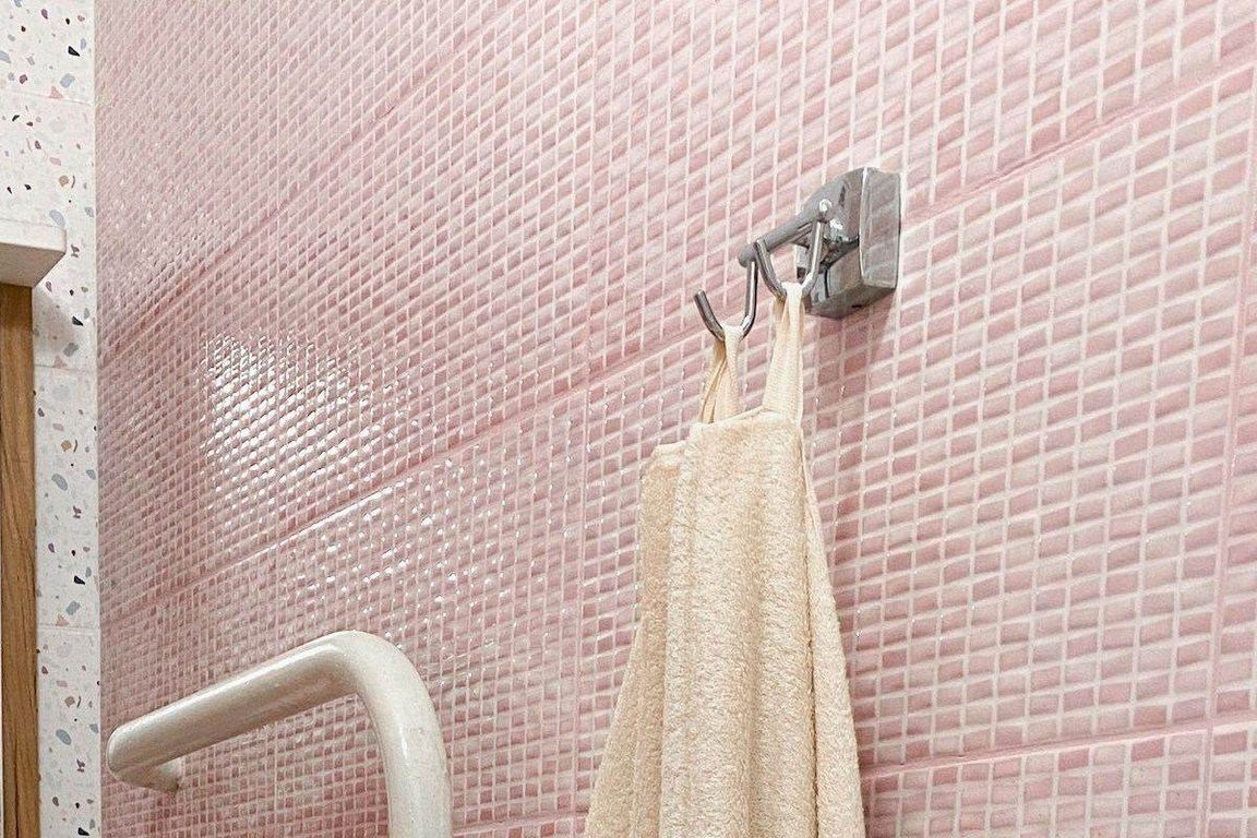 Une serviette de bain beige pend à un crochet métallique à deux branches fixé sur un mur de petits carreaux roses, avec un porte-serviette blanc en bas à gauche.